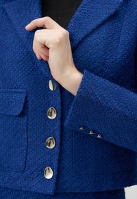 Blue textured fabric vest with a notched collar, large front pockets, and five shiny metallic buttons. Close-up view showcases hand gripping the lapel.