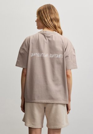 Femme aux longs cheveux portant un T-shirt beige ample avec le texte « PEGADOR » dans le dos et un short beige assorti sur un fond uni.
