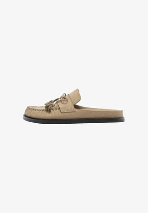 Beige suède loafers zonder achterkant met een strik met kwastjes en een doorgestikte mocassinnaad op een zwarte platte zool, zijaanzicht.