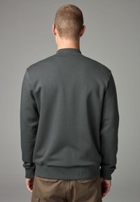 Sweat-shirt gris foncé avec un col rond et des poignets côtelés. Texture lisse, silhouette ajustée et design minimaliste. Pantalon marron visible à la taille.