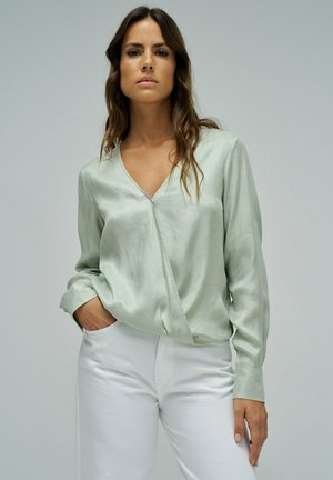 Donna con capelli castani lunghi che indossa una camicia di raso verde chiaro a scollo a V e pantaloni bianchi, in piedi con una mano in tasca davanti a uno sfondo semplice.