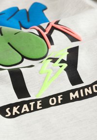 Gråt stof med et grafisk design i blå og grønne farver, sort tekst, lyserøde accenter og et grønt lyn. Teksten lyder "SKATE OF MIND."