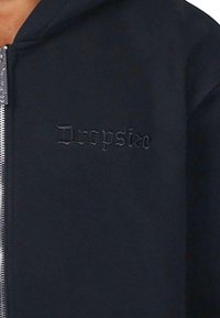 Mustajast zip-up kapuutsiga jakk tekstureeritud kangast. Ees on vastavat äärmust värvi brodüüritud "Dropsize" logo. Hõbedane tõmblukk.