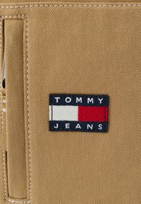 Hnedá tkanina s obdĺžnikovou vyšívanou nášivkou s textom „TOMMY JEANS“ a červenobielo-modrým blokovým dizajnom farieb.