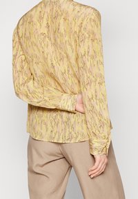 Blusa fluida con maniche lunghe di un giallo chiaro, caratterizzata da un motivo a onde in marrone. Polsini con bottoni e vestibilità ampia. Tessuto morbido.