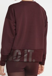 Burgundowa, prążkowana bluza z okrągłym dekoltem, opadającymi ramionami oraz dolną częścią z błyszczącym napisem "ZRÓB TO." na plecach.