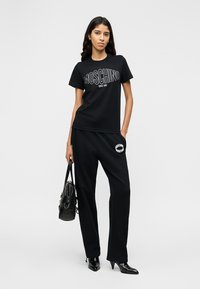 Γυναίκα που φοράει μαύρο t-shirt και παντελόνι Moschino, κρατώντας μια μαύρη τσάντα, στέκεται με το ένα χέρι στην τσέπη.