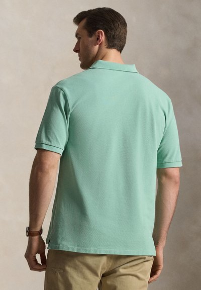 Άνδρας με πράσινο πουκάμισο πόλο με κοντά μανίκια και μπεζ παντελόνι, ορατός από πίσω μπροστά σε μονόχρωμο φόντο.