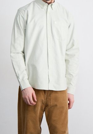 Mann trägt ein langärmliges weißes Hemd mit Knöpfen und Brusttasche sowie braune Cordhosen, steht vor einem schlichten Hintergrund.