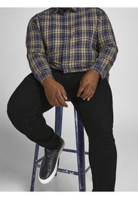 Ruudullinen nappipaita tummansinisestä, vihreästä ja burgundista; mustat istuvat farkut; tummat lenkkarit valkoisilla pohjilla; istuu sinisellä tuolilla.