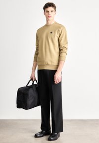 Khaki mikina s okrúhlym výstrihom a malým logom, v kombinácii s čiernymi nohavicami a čiernymi koženými topánkami, držiaca čiernu športovú tašku.