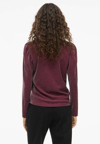 Burgunderfarget langermet topp med subtil glitterstruktur og skrå skuldre, kombinert med svarte bukser, sett bakfra.