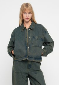 Denim jakna temnega pranja s skrajšanim dizajnom, sprednjim žepom in kontrastnim šivanjem. Zapenjanje na gumbi in ohlapen kroj.