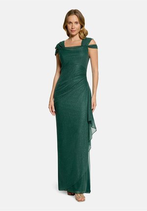 Femme portant une robe de soirée longue verte foncée épaules dénudées avec un léger éclat et des détails froncés, debout devant un fond uni.