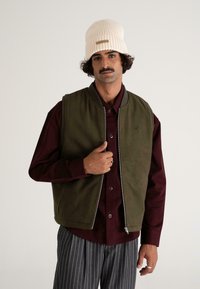 Olive zelena brezrokavnik z zadrgo, oblečen preko burgundske srajce. Vključeni so tudi črtasti hlače in smetana rebrast beanie.