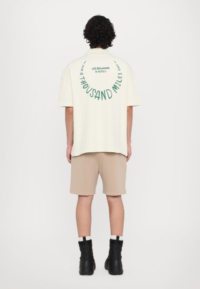 Chemise crème à manches courtes avec un texte vert au dos, associée à un short beige et des bottes noires, offrant une coupe décontractée et une texture douce.