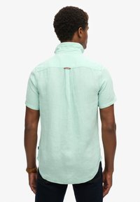 Camicia verde menta a maniche corte in lino. Presenta un colletto ripiegato, una fessura posteriore e un orlo arrotondato, con dettagli di cucitura accentuati.