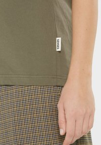Olive green T-shirt with a side label, paired with brown and beige houndstooth patterned skirt. Smooth texture, relaxed fit.