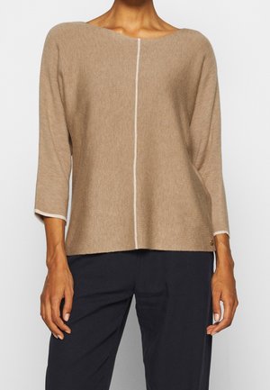 Haut tricoté beige à manches longues avec une couture verticale blanche sur le devant, porté avec un pantalon noir par une personne debout devant un fond uni.