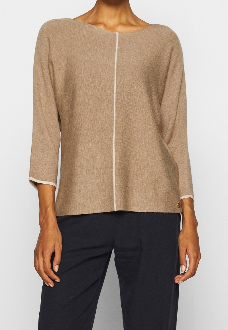 Haut tricoté beige à manches longues avec une couture verticale blanche sur le devant, porté avec un pantalon noir par une personne debout devant un fond uni.
