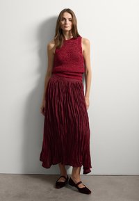 Rotes gestricktes ärmelloses Oberteil mit strukturiertem Muster, kombiniert mit einem langen, fließenden roten Faltenrock. Dunkelrote flache Schuhe runden das Outfit ab.