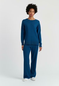 Blauwe gebreide lounge-set met een lange mouwen top en wijde broek. Zachte textuur en minimale naden. G gedragen met witte sportschoenen.