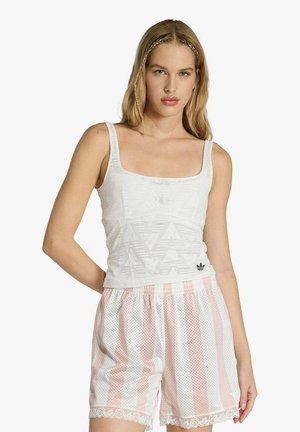 Donna che indossa una canottiera bianca texturizzata e pantaloncini a rete a righe rosa e bianche con bordi in pizzo, in piedi contro uno sfondo semplice.