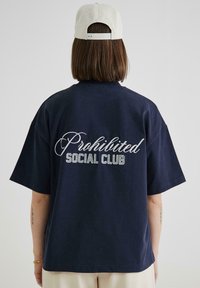 Tamsiai mėlyna per didelė marškinė, pagamintos iš minkštos medvilnės, su baltu siuvinėtų tekstu „Prohibited Social Club“ nugaroje. Trumpomis rankovėmis.