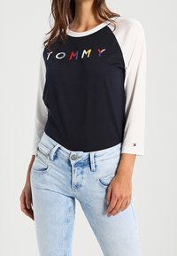 Navy t-shirt with white raglan sleeves and "TOMMY" logo in red, yellow, blue, and white. Paired with light blue jeans.