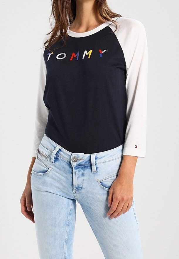 Navy t-shirt with white raglan sleeves and "TOMMY" logo in red, yellow, blue, and white. Paired with light blue jeans.