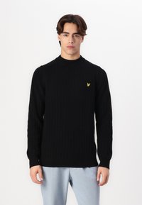 Maglione a costine nero con alta scollatura e maniche lunghe, caratterizzato da un piccolo logo giallo sul petto. Indossato sopra pantaloni blu chiaro.