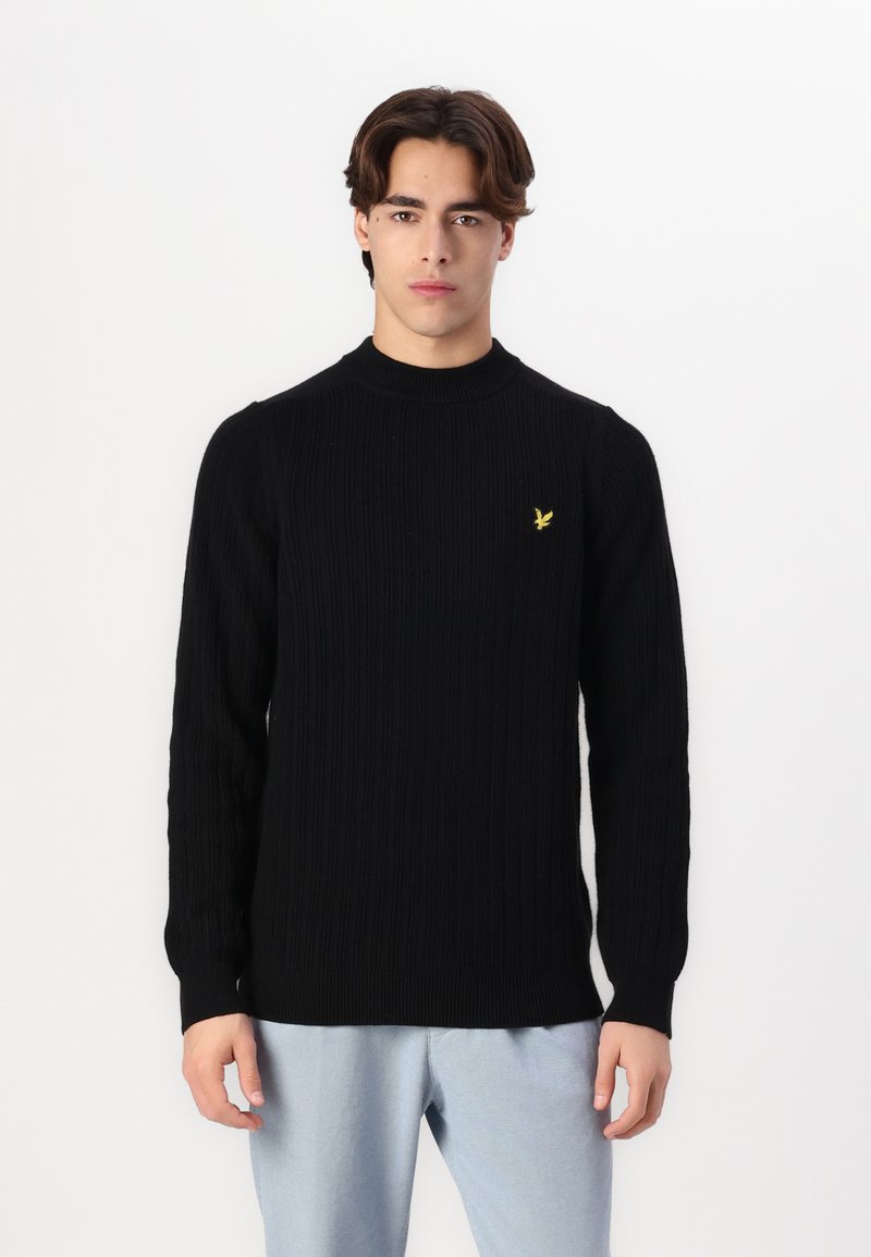 Maglione a costine nero con alta scollatura e maniche lunghe, caratterizzato da un piccolo logo giallo sul petto. Indossato sopra pantaloni blu chiaro.