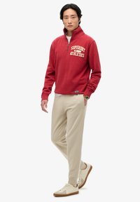 Röd zip-up sweatshirt med texten "Superdry Athletics", beige joggers och krämfärgade sneakers, med en avslappnad passform och mjuk textur.