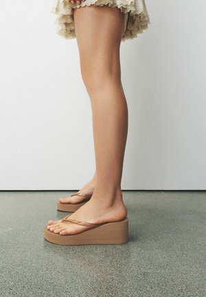 Jalad, millel on beežid kiilkontsaga varbasaapad, õhukeste rihmadega, seisavad hallil põrandal valge seina taustal.