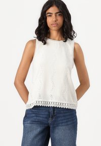 Top blanco sin mangas con tejido texturizado, patrones de encaje ondulados y dobladillo en forma de concha; combinado con jeans azules.