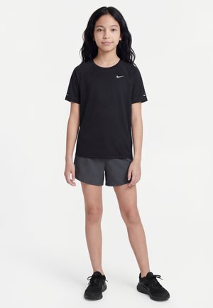 Jeune fille debout, face à l'avant, portant un t-shirt noir Nike, un short gris foncé et des chaussures de sport noires sur un fond blanc.