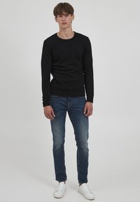 Schwarzer Strickpullover mit rundem Ausschnitt, getragen über blauen Jeans. Weiße Turnschuhe vervollständigen das Outfit. Einfaches, lässiges Design.