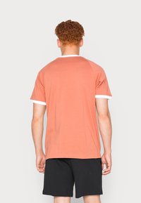 T-shirt s krátkým rukávem v korálové barvě z bavlny s bílými akcenty na límci a lemech rukávů, v kombinaci s černými šortkami, pohled zezadu.