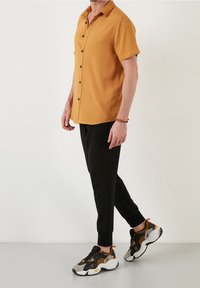 Koszula z krótkim rękawem w kolorze musztardowym, z zapięciem na guziki z przodu, zestawiona z czarnymi, zwężanymi spodniami oraz wielokolorowymi sneakersami z pomarańczowymi akcentami.