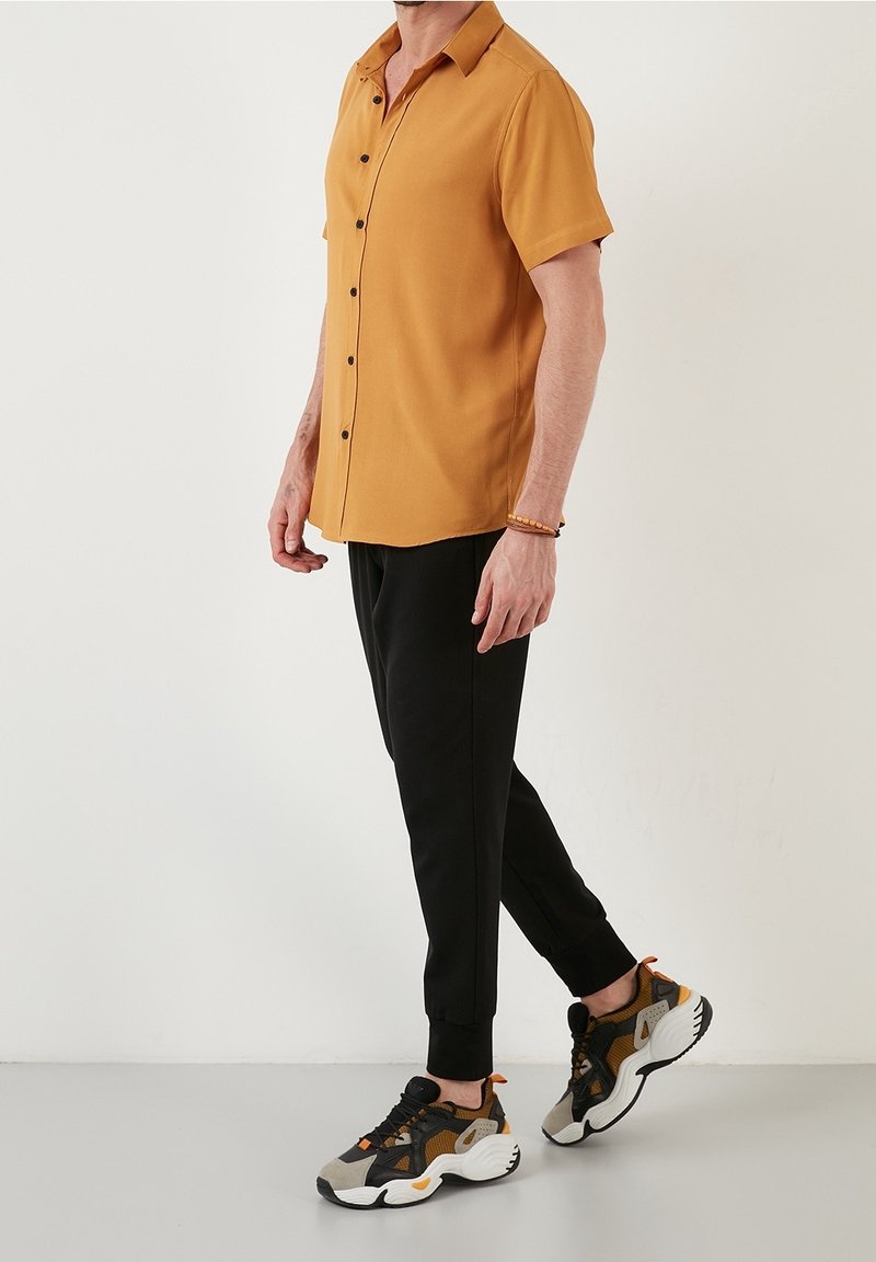 Koszula z krótkim rękawem w kolorze musztardowym, z zapięciem na guziki z przodu, zestawiona z czarnymi, zwężanymi spodniami oraz wielokolorowymi sneakersami z pomarańczowymi akcentami.