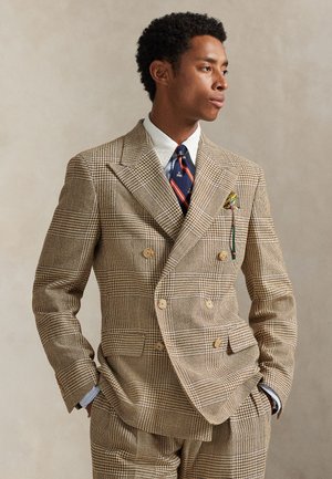 Man draagt een beige dubbelbreasted geruit pak met een wit overhemd, een navy gestreepte stropdas en een patroon zakdoek, handen in de zakken.