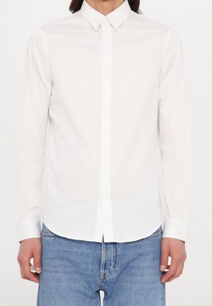 Personne portant une chemise blanche unie à manches longues avec col et un jean bleu en denim, debout devant un fond blanc.