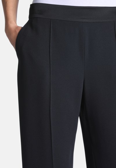 Personne portant un pantalon noir sur mesure avec des poches latérales, une main posée négligemment dans la poche, montrant la texture du tissu et la coupe.
