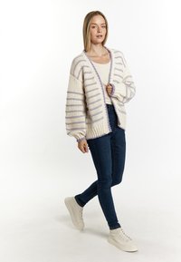 Chunky gebreide cardigan in crème met lila strepen, open voorkant, relaxed fit, geribbelde zoom, gecombineerd met donkere jeans en lichte sneakers.