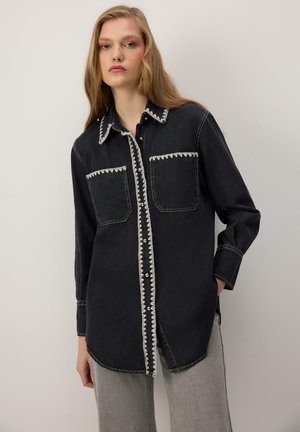 Young woman wearing a black button-up denim shirt with white zigzag trim and light gray pants against a plain neutral background.