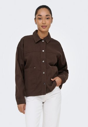 Mujer con chaqueta marrón oscuro abotonada con bolsillos frontales y pantalones blancos, de pie contra un fondo claro y liso, con la mano en el bolsillo.