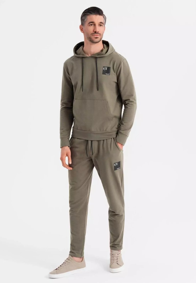 Sudadera verde oliva y joggers a juego, confeccionados en tela suave. Presenta un bolsillo frontal, cordones y un parche con el logo en el pecho y la pierna.