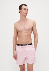 Rosa Badeshorts mit elastischem schwarzen Bund, der das Logo "CALVIN KLEIN" zeigt. Leichter Stoff, Kordelzugverschluss und eine Länge über dem Knie.