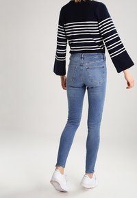 Persona con un suéter a rayas azul marino y blanco, jeans skinny azul claro y zapatillas blancas, caminando hacia atrás sobre un fondo simple.