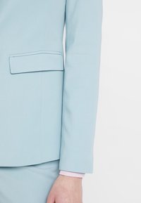 Blazer bleu clair avec une coupe ajustée, doté d'une poche poitrine simple et d'une texture de tissu lisse ; poignet côtelé rose visible.